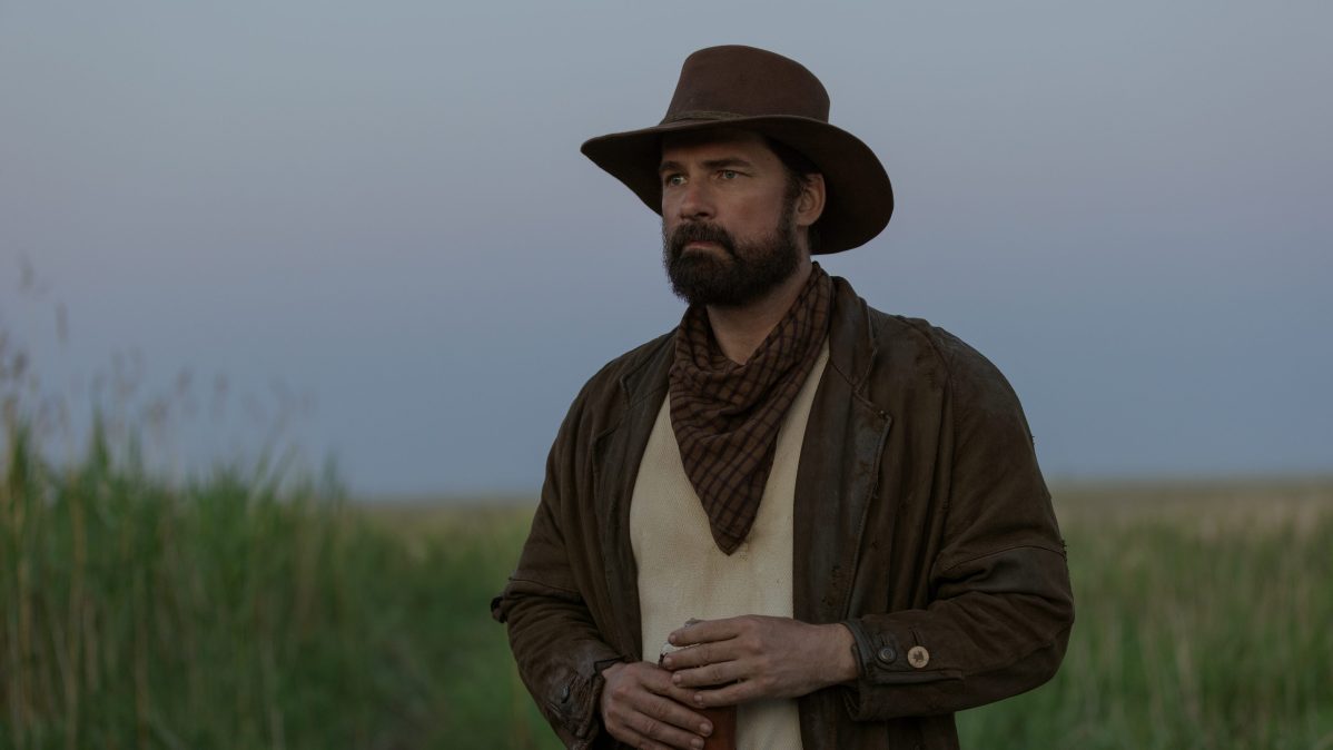 "Man dressed as a cowboy with a brown hat and bandana stands in a grassy field at dusk, looking into the distance. The sky is clear and the atmosphere is calm, evoking a western or frontier setting."