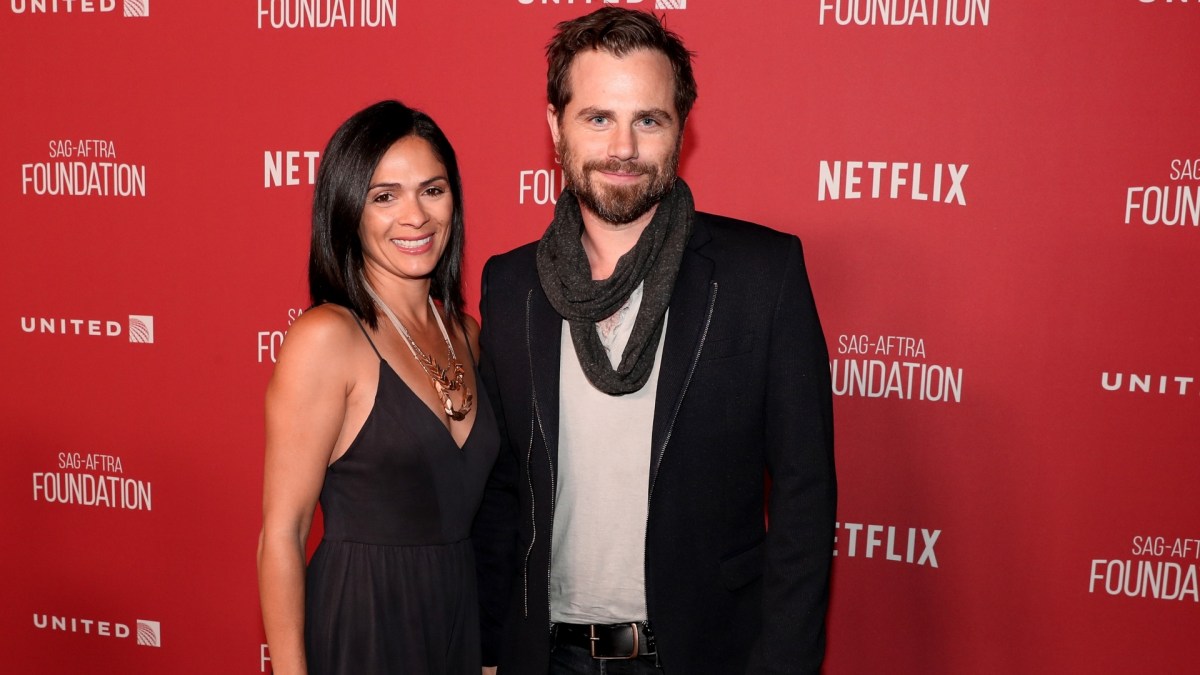 Alexandra Barreto (L) and Rider Strong pose in black outfits in front of a red Netflix background.