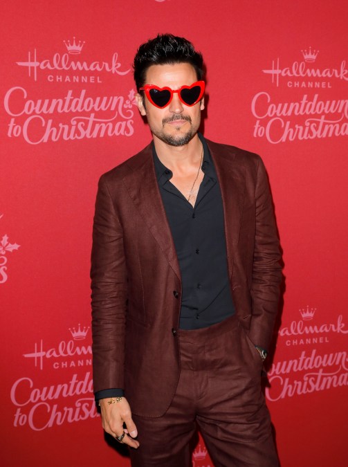 Tyler Hynes smiles in brown suit, black shirt, and red heart sunglasses