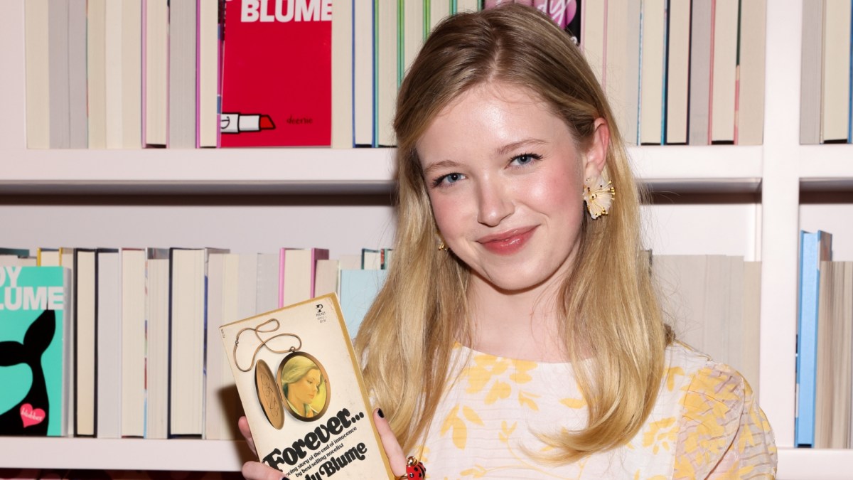 Shay Rudolph smiles in front of a bookshelf, holding the Judy Blume book, "Forever."