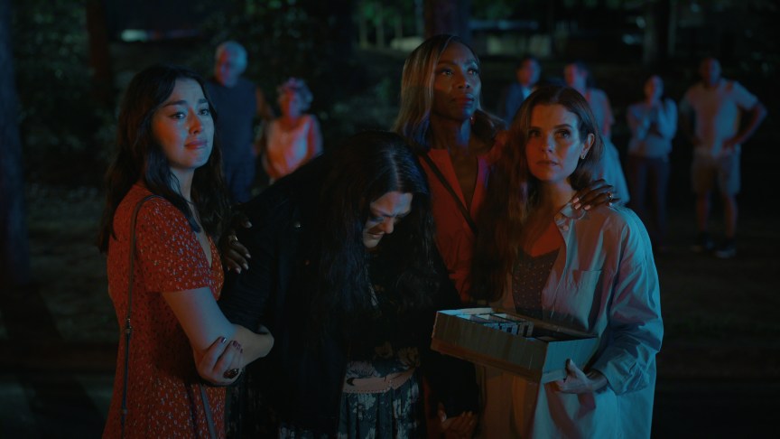"Four young women standing together at night, two comforting another who is hunched over, with concerned onlookers and trees in the background. The scene is lit with blue and red lights, suggesting a dramatic or tense moment outdoors."