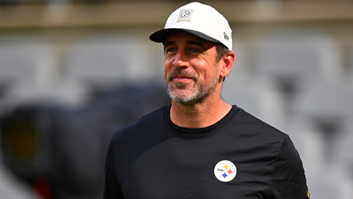 Aaron Rodgers stands on a football field wearing a Pittsburgh Steelers hat.